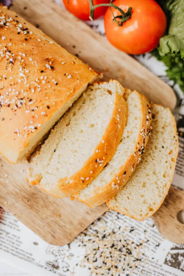 A loaf of bread, sprinkled with homemade everything bagel seasoning, rests partially sliced on a wooden cutting board. Fresh tomatoes and greens add vibrant color in the background, while crumbs and seeds are scattered around aesthetically.