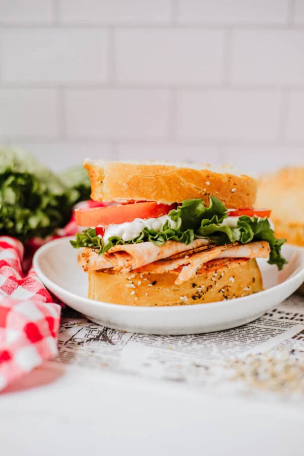 A delicious sandwich on a white plate, featuring sliced turkey, fresh lettuce, tomato, and poppy seed bread sprinkled with homemade everything bagel seasoning. A red and white checkered cloth is nearby, with greens blurred in the background. The setting is a bright kitchen counter.