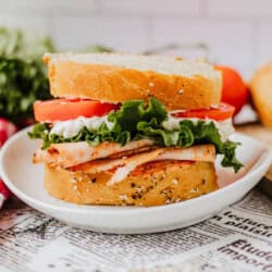 A close-up of a turkey sandwich on a white plate. It features slices of turkey, lettuce, and tomato with mayonnaise between two slices of seeded bread sprinkled with homemade everything bagel seasoning. The background shows a blurred newspaper, cherry tomatoes, and lettuce.