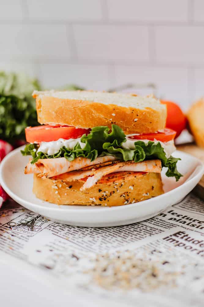 A close-up of a turkey sandwich on a white plate. It features slices of turkey, lettuce, and tomato with mayonnaise between two slices of seeded bread sprinkled with homemade everything bagel seasoning. The background shows a blurred newspaper, cherry tomatoes, and lettuce.