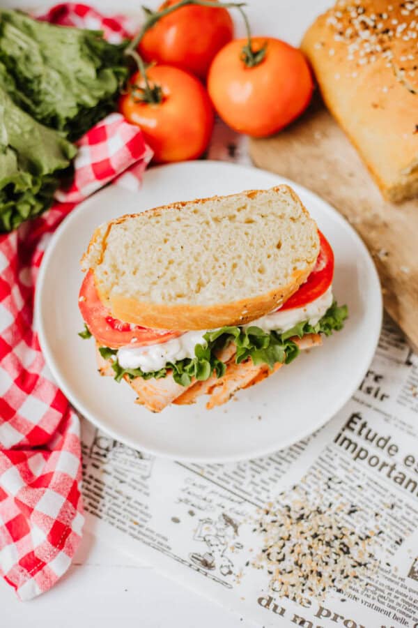 A sandwich on a white plate sits atop a newspaper, featuring lettuce, tomato, and cheese between two slices of sesame bread sprinkled with homemade everything bagel seasoning. Surrounding the plate are fresh tomatoes, a head of lettuce, a bread loaf, and a red gingham napkin.