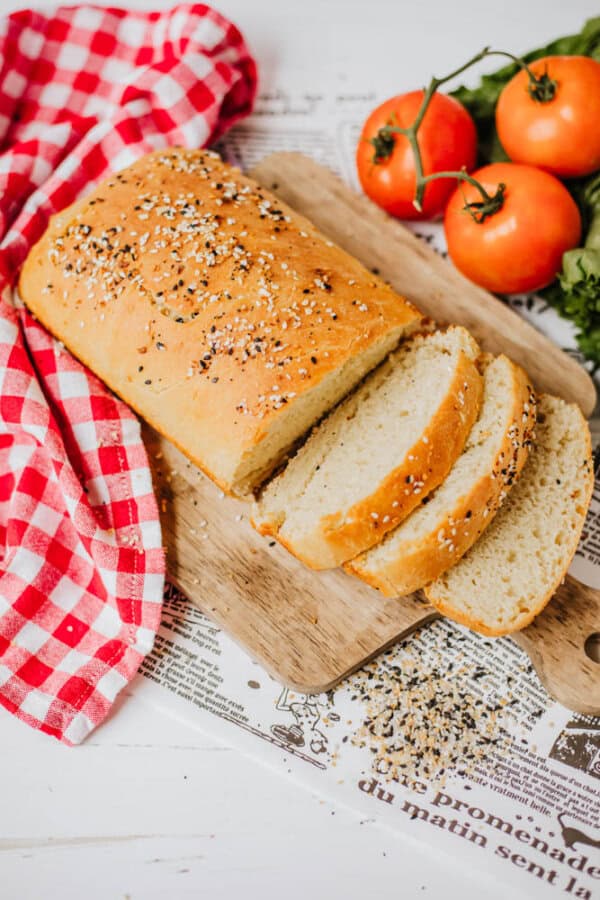 A sliced loaf of homemade bread sprinkled with everything bagel seasoning rests on a wooden cutting board. Next to it, a red and white checkered cloth complements the fresh tomatoes on the vine nearby.