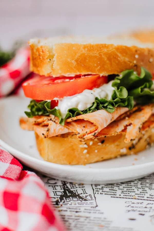 A close-up of a turkey sandwich on a white plate, featuring slices of turkey, lettuce, tomato, and a dollop of mayonnaise between two slices of bread dusted with homemade everything bagel seasoning. A red and white checkered napkin is partially visible to the side.
