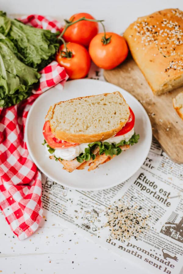 A sandwich filled with lettuce, tomato, and a slice of chicken is placed on a white plate. Sprinkled with homemade everything bagel seasoning for an extra kick, the scene is complete with fresh tomatoes, lettuce, a checkered red napkin, and bread on a wooden board atop a newspaper sheet.