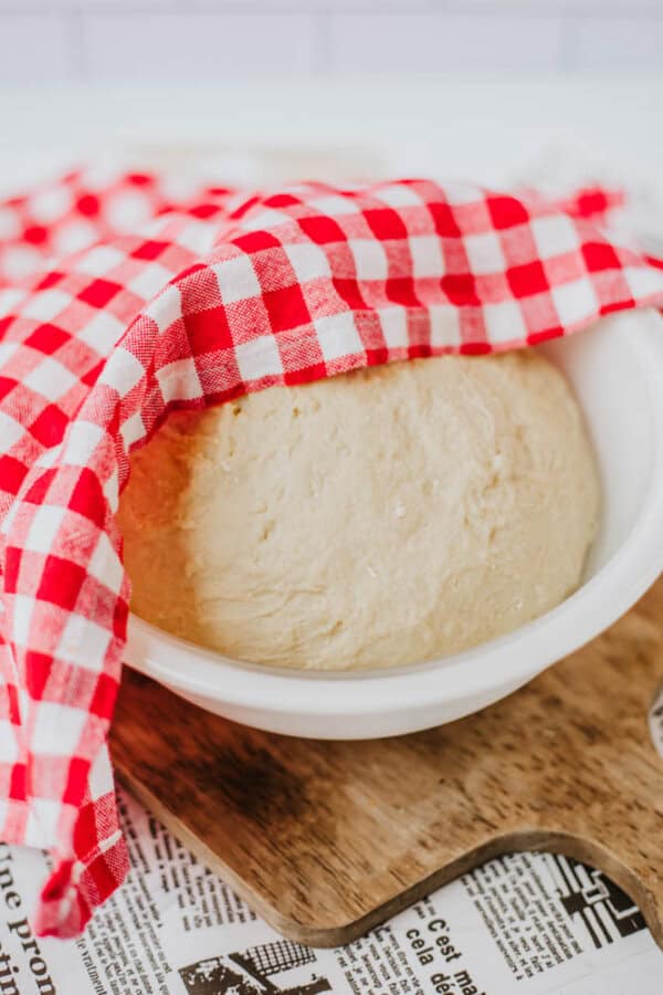 A bowl of dough sprinkled with homemade everything bagel seasoning is covered with a red and white checkered cloth, resting on a wooden board atop newspaper.