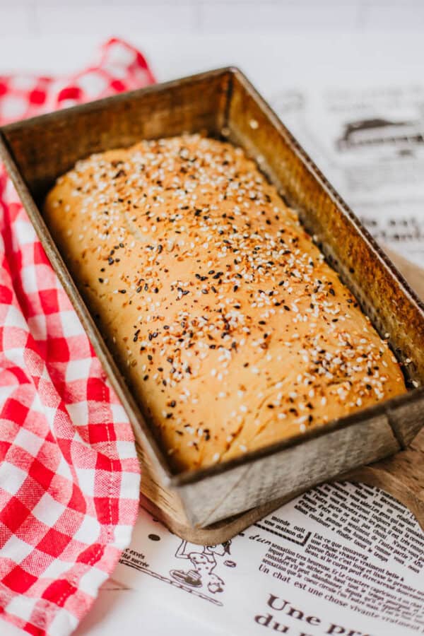 A loaf of freshly baked bread sprinkled with homemade everything bagel seasoning rests in a metal pan on the kitchen countertop. A red and white checkered cloth lies nearby, with printed text and illustrations beneath it.