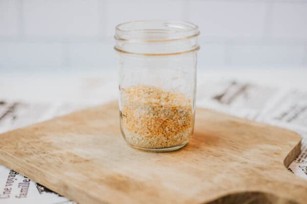 A glass jar filled with homemade everything bagel seasoning, featuring dried minced garlic, sits on a wooden cutting board. The background is a white tiled wall, and there are newspapers underneath the board.