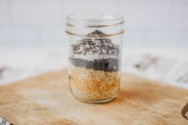 A glass jar brimming with layers of dry ingredients like homemade everything bagel seasoning, black seeds, white grains, and yellow flakes sits on a wooden surface against a blurred background.
