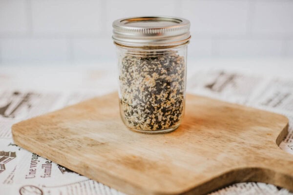 A mason jar filled with homemade everything bagel seasoning, a delightful blend of dried seeds and spices, sits on a wooden cutting board. The background features a white tiled wall and a newspaper-covered surface, adding to its rustic charm.