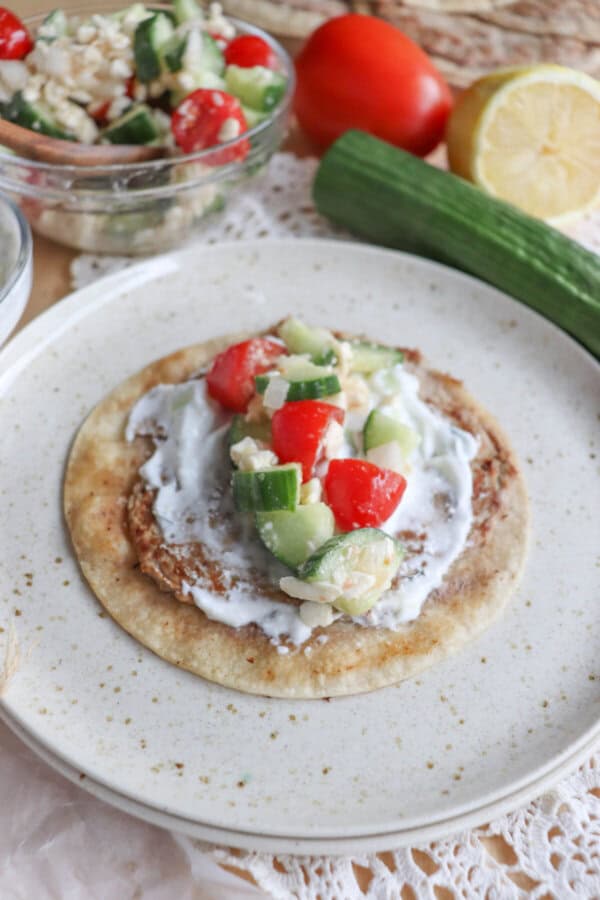 A plate with a flatbread, reminiscent of Greek cuisine, topped with a creamy spread and chunky salad made of cucumber, tomatoes, and grains. Nearby are fresh ingredients ready to create fusion dishes like turkey smash tacos on a lace tablecloth.
