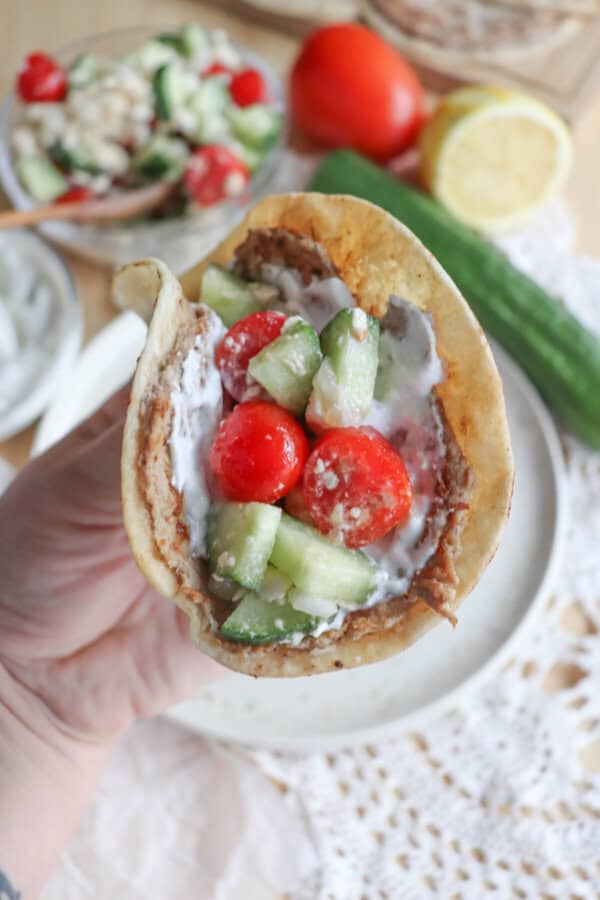 A hand holds a pita filled with creamy sauce, sliced cucumbers, and cherry tomatoes over a layer resembling beans. This Greek-inspired creation brings to mind turkey smash tacos. In the background lie a halved lemon, cucumber, onion slices, and a bowl of salad on a lace-covered table.