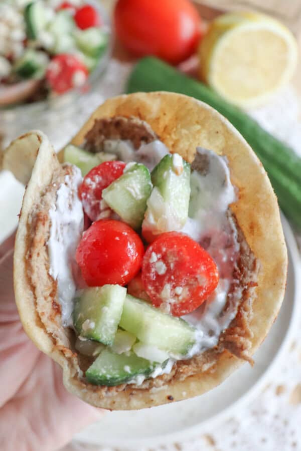A close-up of a Greek pita wrap filled with sliced cucumbers, cherry tomatoes, and creamy sauce. The pita is held in hand, while a lemon, whole cucumber, and more salad are visible in the background. Its reminiscent of vibrant turkey smash tacos but with a Mediterranean twist.