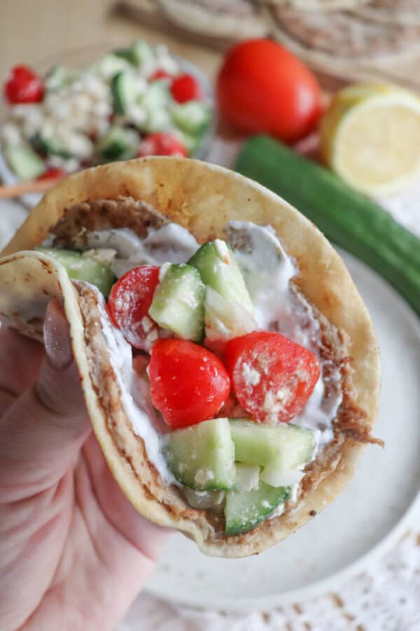 A hand holding a Greek-inspired pita filled with sliced cucumbers, cherry tomatoes, and feta cheese drizzled with creamy sauce. A cucumber, tomato, and lemon are blurred in the background, adding a fresh vibe to these vibrant flavors.