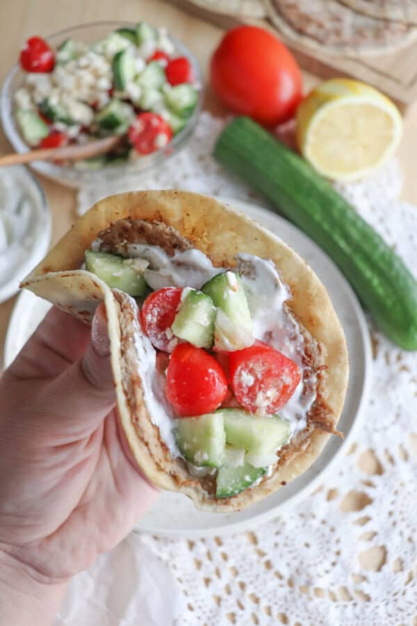 A person holds a pita filled with creamy spread, diced cucumber, and cherry tomatoes, reminiscent of Greek flavors. In the background, fresh ingredients like cucumber, tomato, and a salad bowl are set on a lace tablecloth.