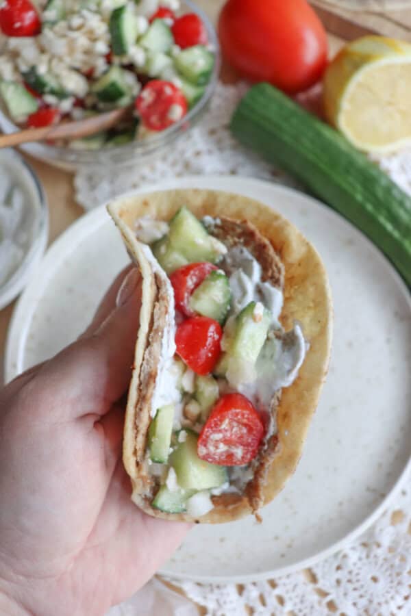 A hand holds a pita filled with sliced cucumber, cherry tomatoes, lettuce, and a creamy sauce reminiscent of Greek flavors. In the background, fresh ingredients like cucumber, tomato, and lemon rest on a lace tablecloth. It’s a refreshing twist akin to a delicious turkey smash taco experience.