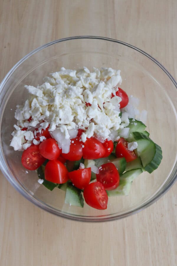 A glass bowl filled with cherry tomatoes, cucumber slices, and chopped onions, all topped with crumbled feta cheese, evokes a Greek feast. Set on a wooden surface, it complements the flavors of Greek turkey smash tacos beautifully.