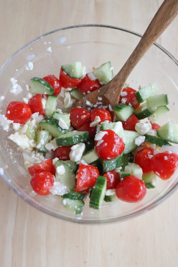 A clear bowl on a wooden surface is filled with a Greek-inspired salad featuring diced cucumbers, cherry tomatoes, and crumbled feta cheese. A wooden spoon rests inside the bowl, blending the vibrant ingredients together like a culinary nod to turkey smash tacos.