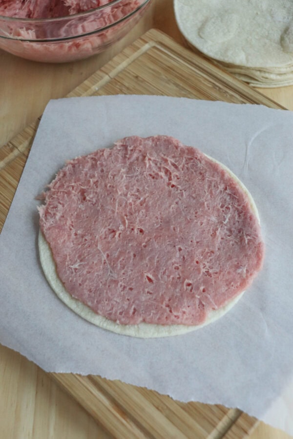 A thin layer of pink, shredded turkey meat spreads over a round, white tortilla on a sheet of parchment paper. In the background, a bowl contains more shredded meat for these Greek-inspired smash tacos, and there are additional tortillas waiting on a wooden surface.
