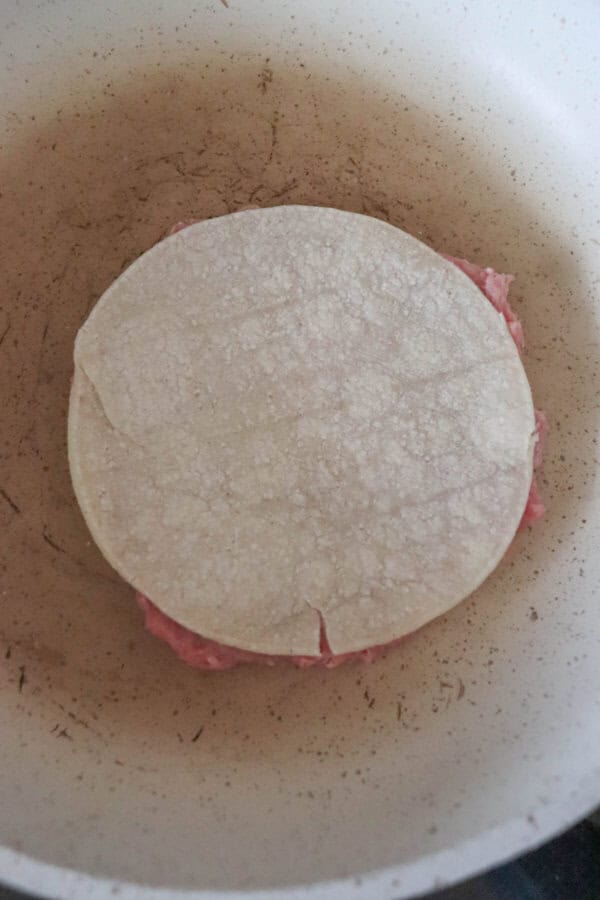 A corn tortilla is placed in a frying pan over raw minced turkey, kickstarting the process of crafting a delicious taco or similar dish. The pan, seasoned with experience, shows slight discoloration as it brings these Greek-inspired smash tacos to life.