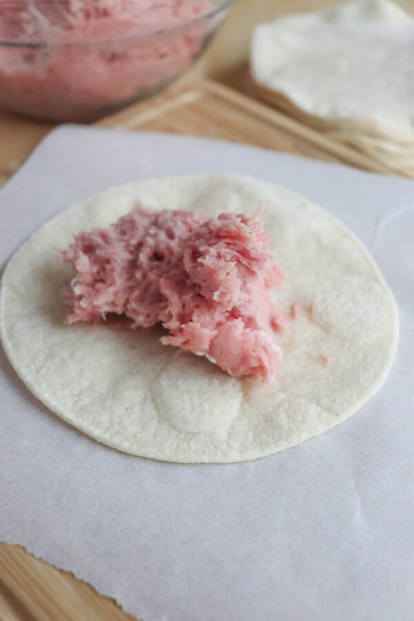 A corn tortilla rests on parchment paper with a scoop of pink filling in the center, reminiscent of Greek turkey smash tacos in preparation. In the background, a bowl brimming with more filling and extra tortillas awaits, ready to complete this delectable creation.