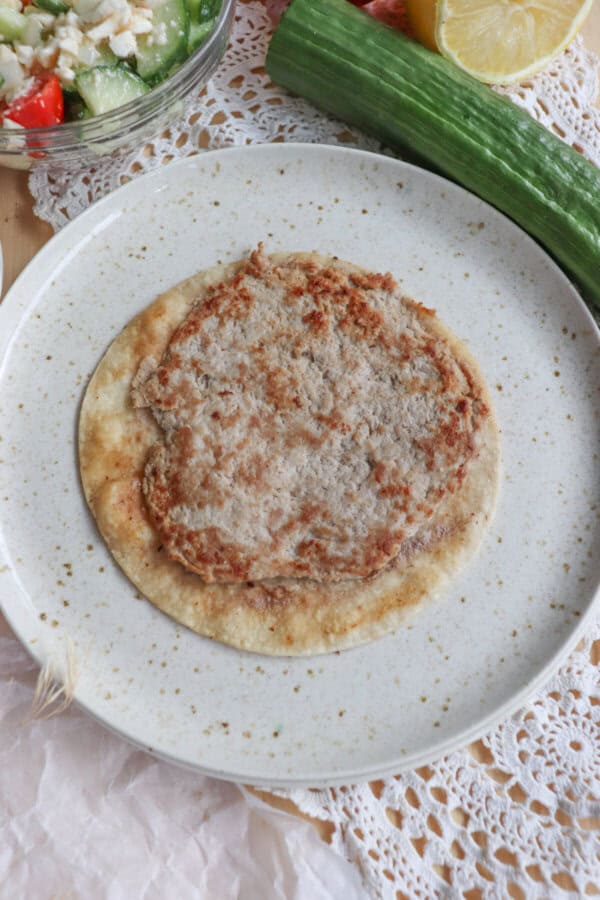 A Greek-inspired flatbread topped with minced meat sits on a speckled plate. Beside it lie a cucumber, half a lemon, and a vibrant salad of cucumbers, tomatoes, and feta cheese atop a lace-textured surface.