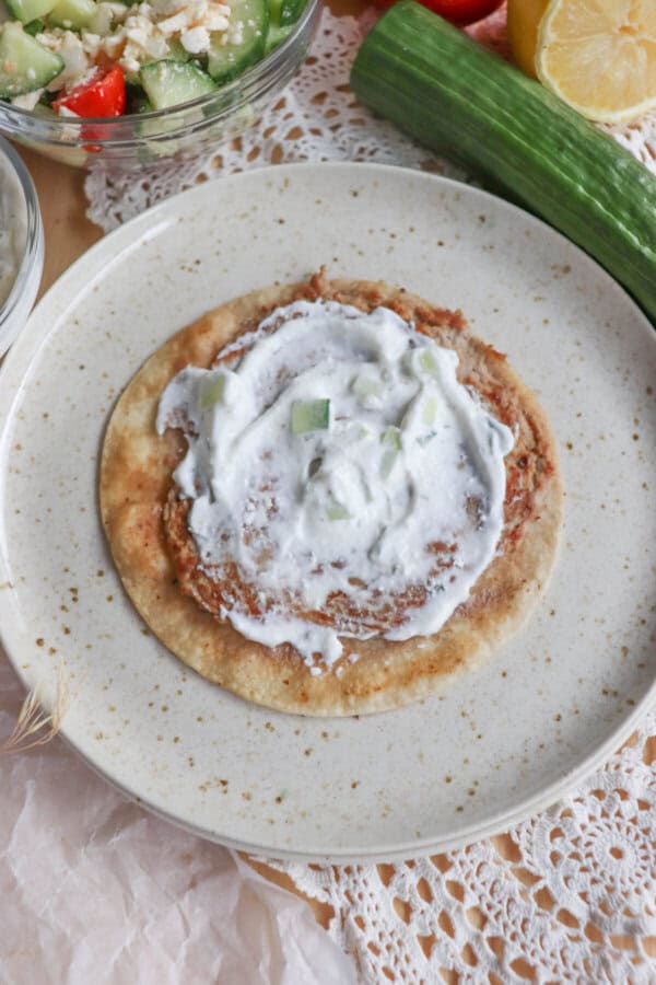 A Greek-inspired flatbread topped with shredded meat and tzatziki sauce sits on a speckled plate. A cucumber and lemon are nearby, hinting at fresh turkey flavors. In the background, theres a bowl of mixed salad with tomatoes, cucumber, and cheese on a lace tablecloth.