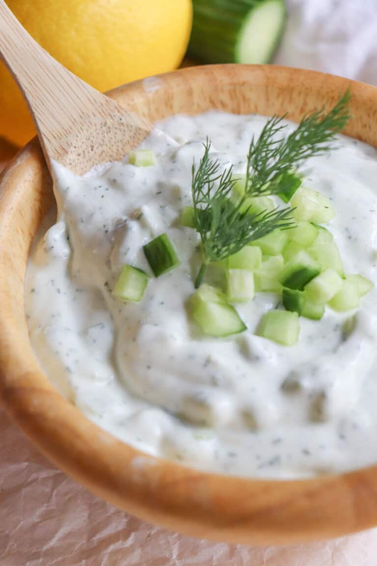 A wooden bowl filled with creamy, high-protein tzatziki sauce is garnished with diced cucumber and a sprig of dill. A wooden spoon rests in the bowl, with lemon and cucumber slices softly blurred in the background.