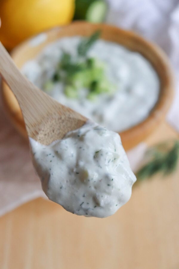 A wooden spoon holds a dollop of creamy, high-protein tzatziki sauce with visible herbs and cucumber bits. In the background, a bowl filled with more tzatziki and a lemon can be seen slightly blurred.
