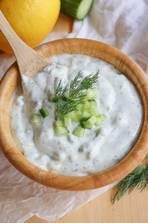 A wooden bowl of creamy, high-protein tzatziki sauce garnished with chopped cucumber and fresh dill, with a wooden spoon resting inside. A lemon and a cucumber lie in the background on a white cloth.