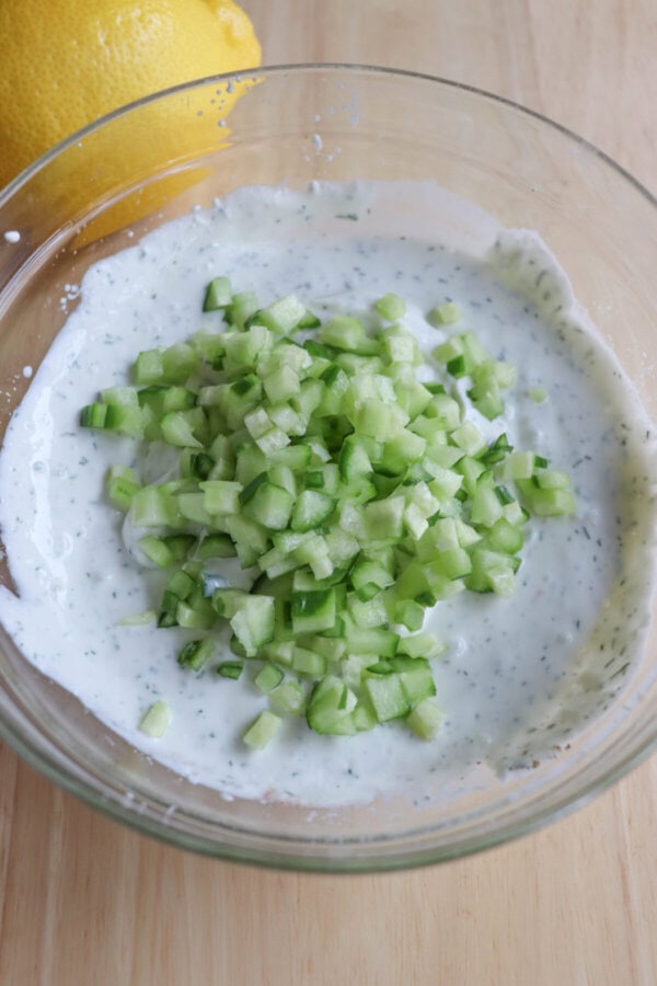 A clear glass bowl filled with creamy, high-protein tzatziki sauce topped with diced cucumber. A whole lemon is partially visible in the background on a wooden surface.
