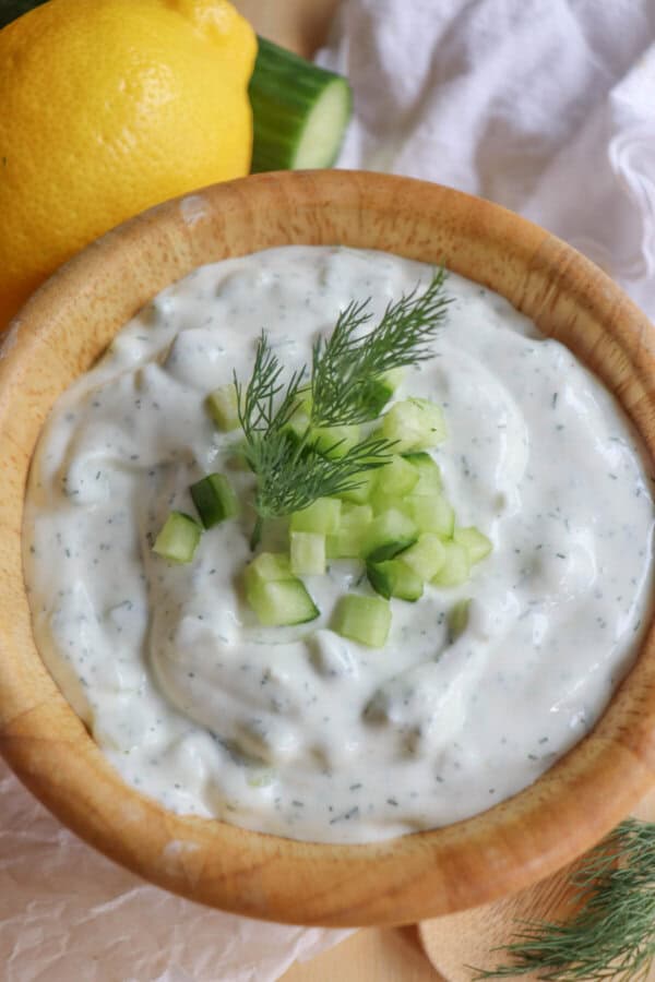 A wooden bowl filled with creamy, high-protein tzatziki sauce garnished with chopped cucumber and dill. A lemon and cucumber are nearby on a light cloth background.