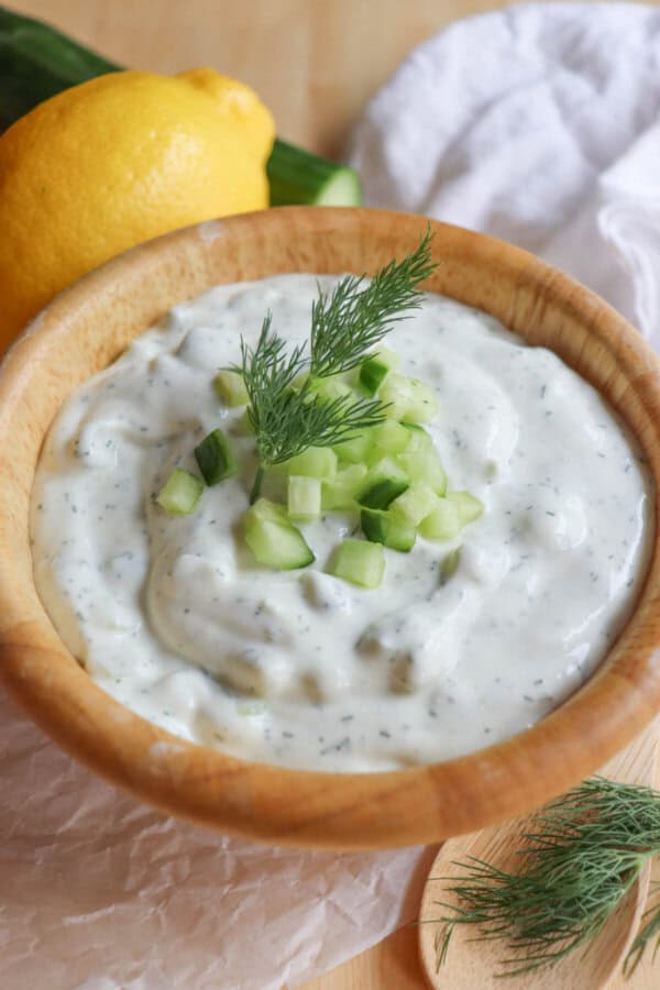 A wooden bowl filled with creamy, high-protein tzatziki sauce is garnished with chopped cucumber and a sprig of dill. Nearby, a lemon, cucumber, and sprig of dill accompany a wooden spoon on a white cloth.