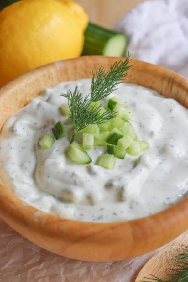 A wooden bowl brimming with high protein tzatziki sauce, garnished with fresh cucumber pieces and a sprig of dill. A lemon and part of a cucumber rest in the background on a rustic wooden surface, adding to the scenes vibrant freshness.