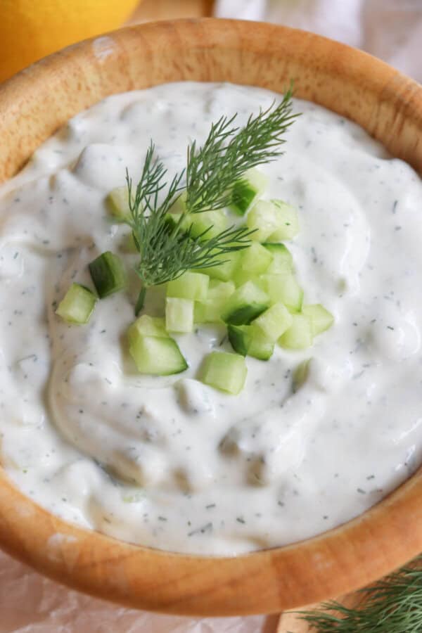 A wooden bowl filled with creamy, high-protein tzatziki sauce, garnished with fresh cucumber cubes and a sprig of dill. The sauce boasts a smooth texture, showcasing a rich mix of yogurt, cucumber, garlic, and herbs.