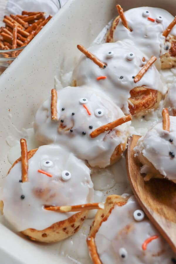 A baking dish with cinnamon rolls frosted to look like snowmen, complete with candy eyes and pretzel stick arms. A small bowl of pretzel sticks is in the background.