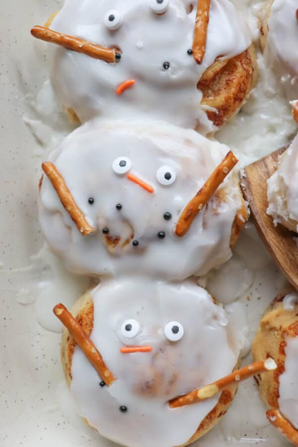 Three frosted pastries decorated to look like snowmen, featuring candy eyes, orange sprinkle noses, and pretzel stick arms. The treats are arranged in a tray, with one on a wooden spatula.