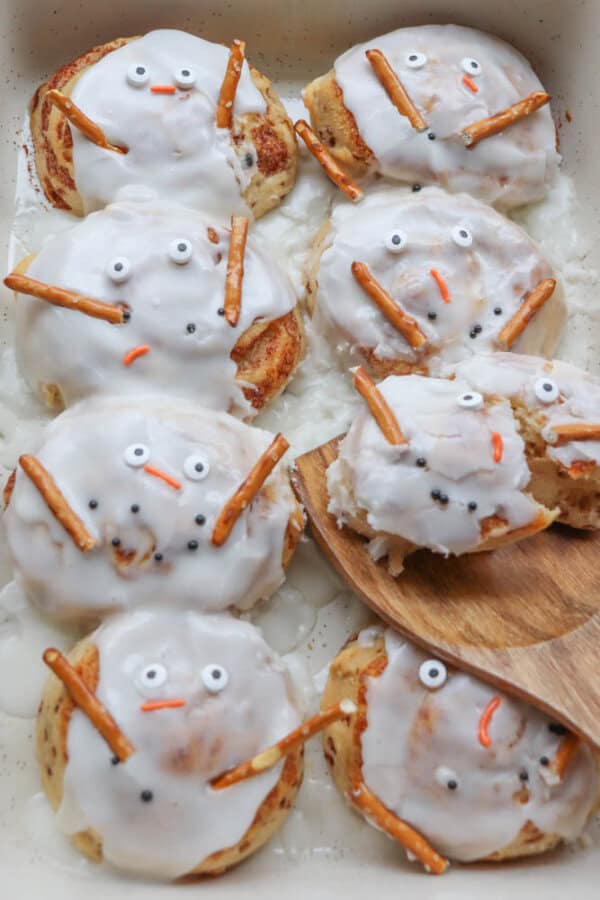 Cinnamon rolls decorated to look like snowmen with white icing, candy eyes, pretzel stick arms, and orange candy noses, arranged in a baking dish. A wooden spatula holds one cinnamon roll.