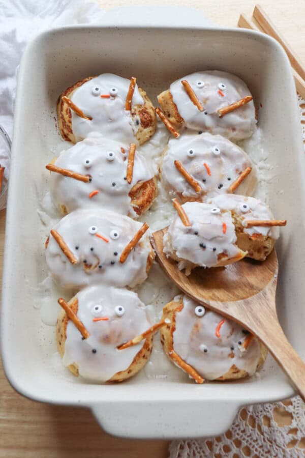 A baking dish filled with cinnamon rolls decorated to resemble snowmen. Each roll is frosted with white icing and features pretzel stick arms, candy eyes, and orange candy for noses, creating cute snowman faces. Wooden spoon on the side.