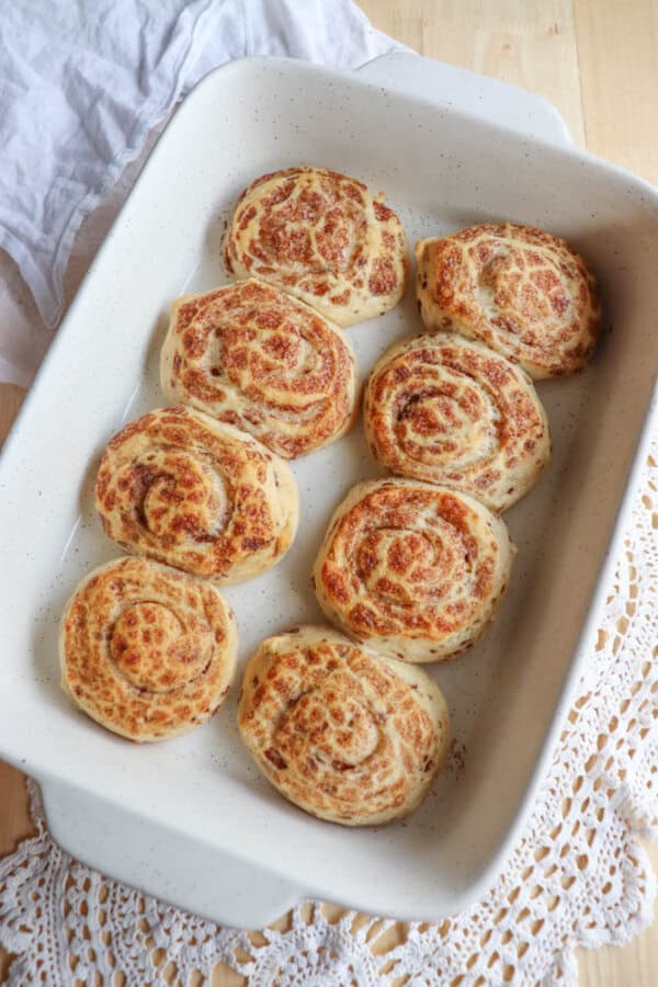 A baking dish with eight freshly baked cinnamon rolls, arranged in two rows, on a light wooden table. A white cloth is partially visible in the background.