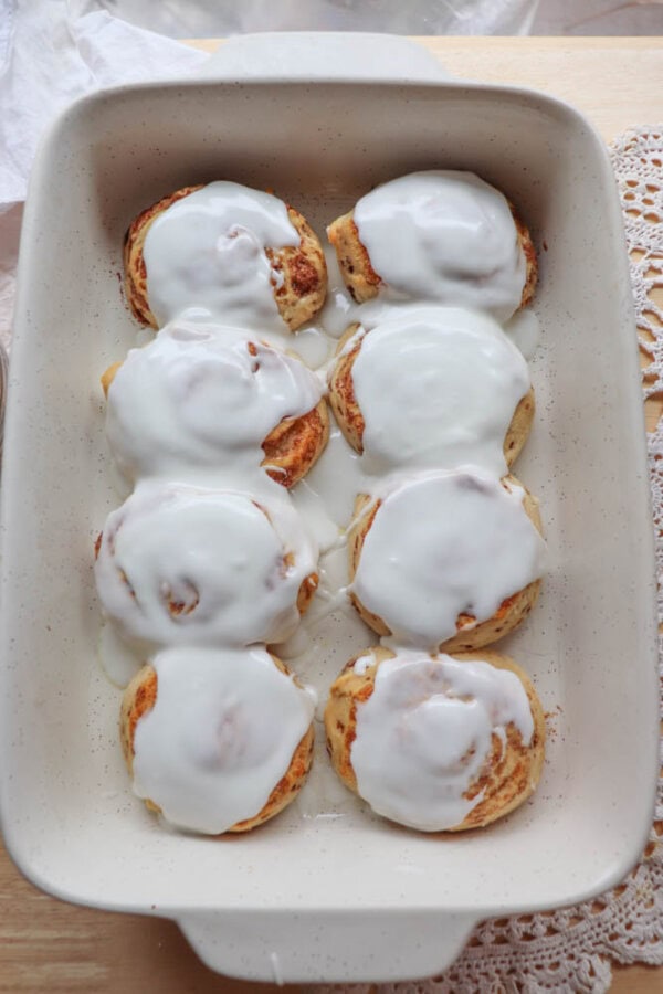 A rectangular baking dish filled with eight frosted cinnamon rolls. The rolls are evenly spaced and covered with a generous layer of white icing. The dish is placed on a wooden surface with a lace cloth partially visible on the side.