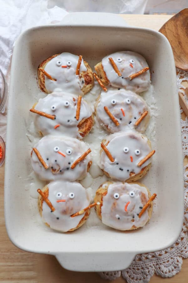 A baking dish with eight snowman-themed cinnamon rolls topped with white icing. Each has candy eyes, an orange candy nose, and pretzel stick arms, arranged to resemble snowmen faces. The dish sits on a wooden surface with a doily nearby.
