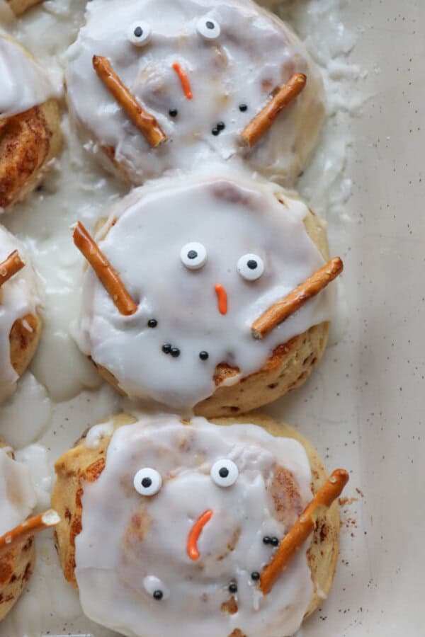 Cinnamon rolls decorated as snowmen, with white icing, candy eyes, pretzel arms, and orange candy noses, arranged in a baking pan.