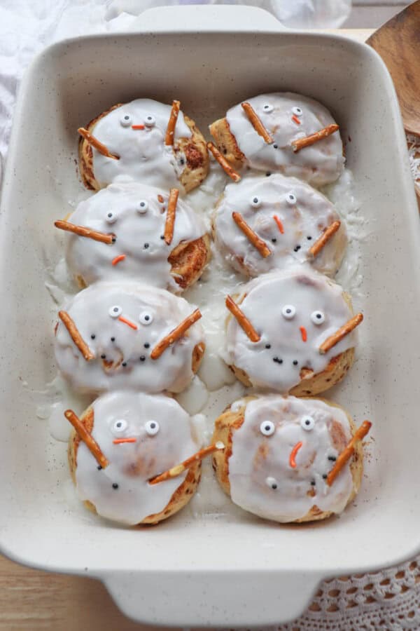 A baking dish filled with cinnamon rolls decorated to look like snowmen. Each roll is covered with white icing and has candy eyes, a carrot-shaped nose, and pretzel sticks for arms.