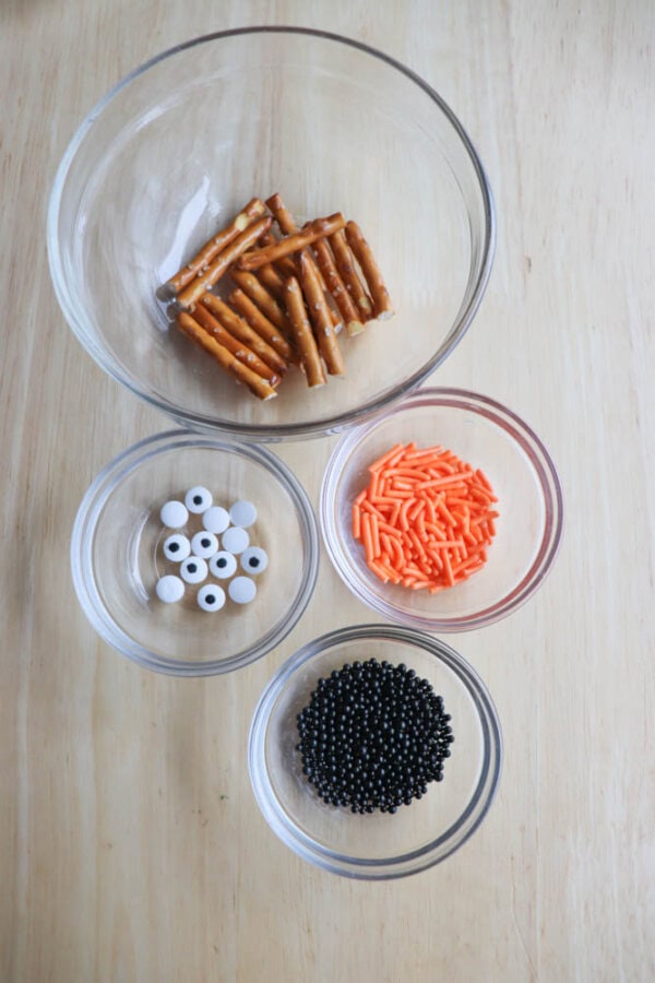 Four clear bowls on a wooden surface contain pretzel sticks, orange sprinkles, black round sprinkles, and white candy eyes.