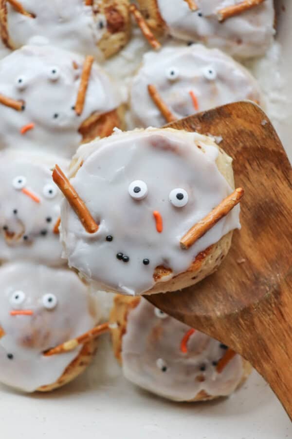 Cinnamon rolls decorated as snowman faces with white icing, candy eyes, an orange candy nose, and pretzel stick arms on a wooden spatula.