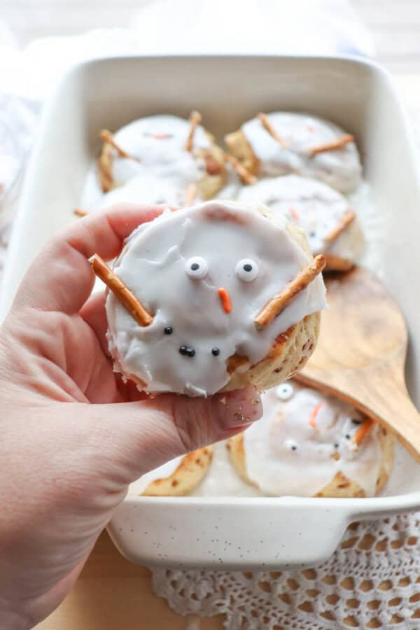 A hand holding a cinnamon roll decorated to look like a snowman, with white icing, pretzel stick arms, candy eyes, and a small orange candy for the nose. More snowman-themed rolls are in a baking dish in the background.