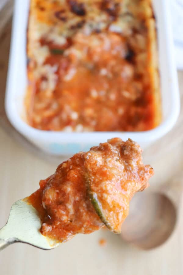 A fork holds a piece of zucchini lasagna, rich with savory tomato sauce, in front of a baking dish filled with more. The wooden surface adds rustic charm to this delightful single serving treat.