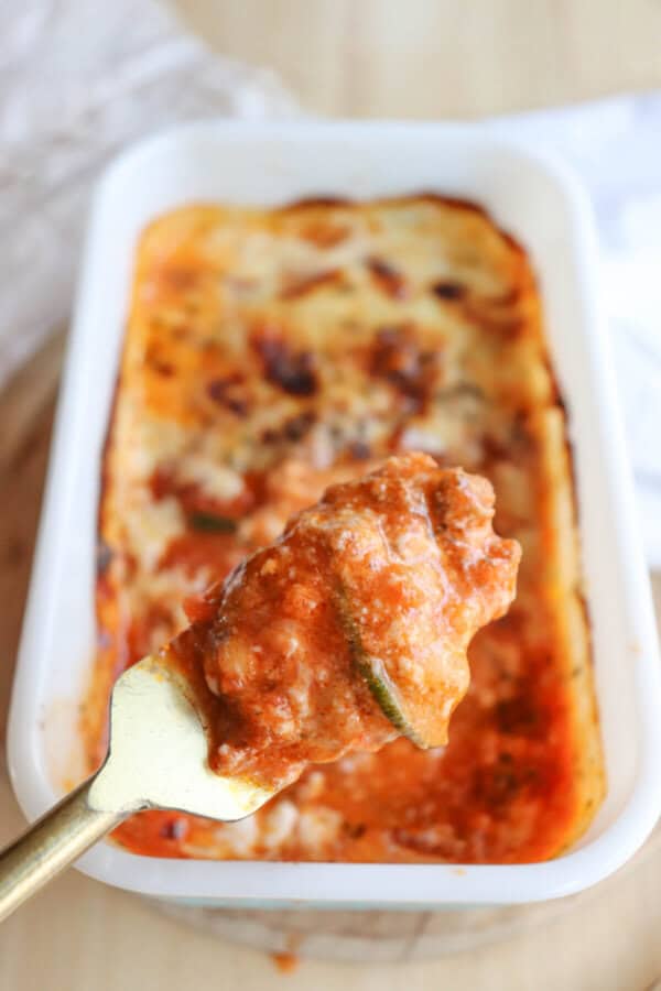 A close-up of a fork holding a piece of zucchini lasagna reveals layers of pasta, tomato sauce, and melted cheese. The background shows a rectangular white baking dish with the remaining single serving on a wooden surface.