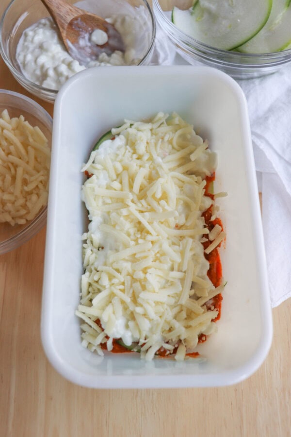 A white baking dish features a delightful single serving zucchini lasagna, layered with red sauce and topped with shredded cheese. Surrounding it are small bowls brimming with extra cheese, cottage cheese, and sliced green apples. A wooden spoon rests in one bowl.