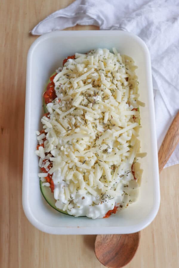 A white baking dish holds a single serving zucchini lasagna, layered with zucchini, red sauce, grated cheese, and herbs. Ready for baking, its set against a light wooden surface with a wooden spoon and a white cloth nearby.